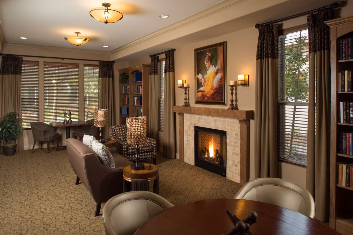 Warm, well-lit common living room with a lit fireplace, sofas and armchairs, bookshelves, and large windows with blinds.