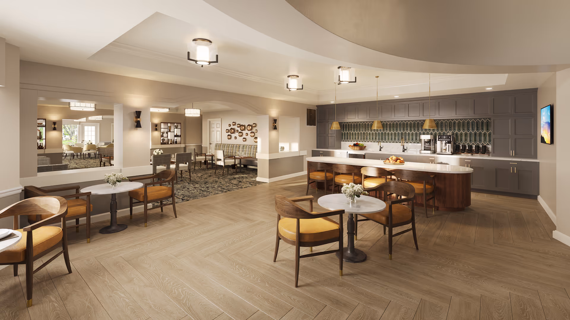 A spacious and well-lit common area in an assisted living facility featuring a modern kitchen with gray cabinets, a white countertop island with wooden bar stools, and pendant lights. The room includes small round tables with chairs, decorative wall art, and a large mirror reflecting another seating area with multiple chairs and tables.