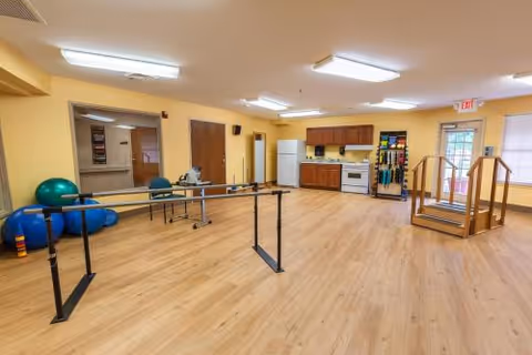 A spacious rehabilitation room with wooden flooring, featuring parallel bars for walking exercises, exercise balls, a small kitchen area with a refrigerator, stove, and cabinets, and a wooden staircase with handrails for physical therapy. The walls are painted yellow, and the room is well-lit with ceiling lights.