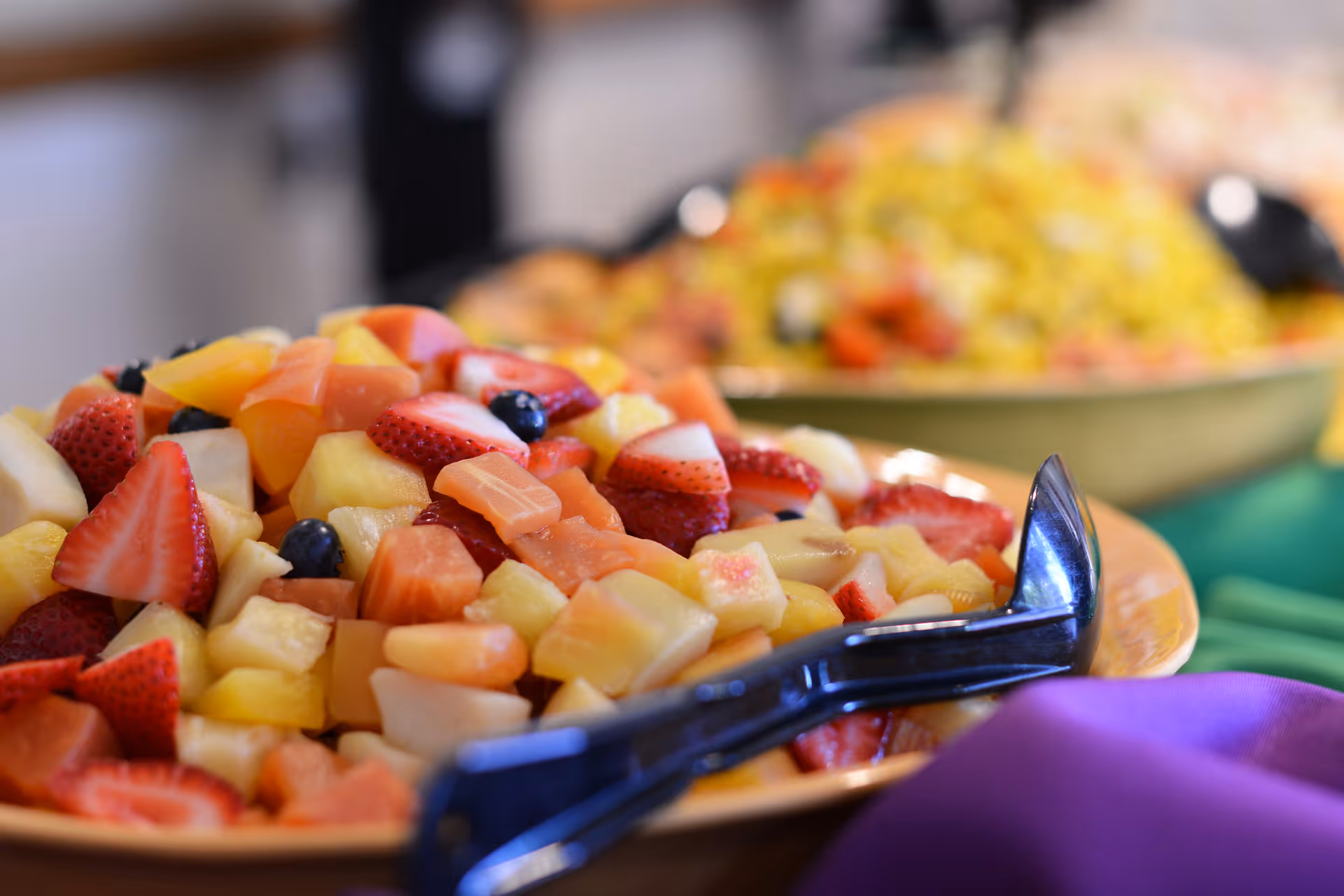 Close-up view of a plate filled with a colorful fruit salad including strawberries, pineapple, blueberries, and other mixed fruits with a serving utensil on the side. In the background, there is another dish with a yellow rice salad and a green tablecloth underneath.