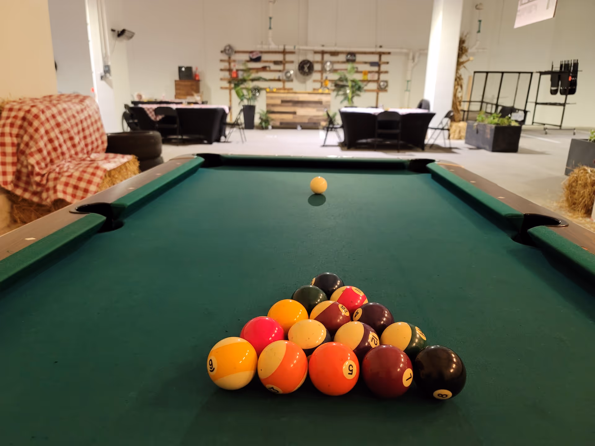 View down a pool table with racked billiard balls in a large indoor recreation room with tables and seating in the background.