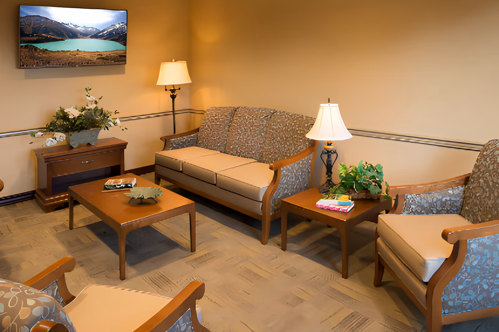 A cozy living room with a patterned three-seat sofa, two matching armchairs, a wooden coffee table with decorative items, two side tables with lamps and plants, and a wall-mounted TV displaying a scenic mountain and lake view.