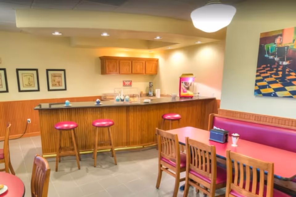 A cozy dining area with a wooden counter and three red cushioned bar stools. There are tables with wooden chairs and red cushioned bench seating. The counter has a popcorn machine, a coffee maker, and some small decorative items. The walls are light yellow with framed pictures and a colorful painting. The floor is tiled and the ceiling has recessed lighting and a hanging light fixture.