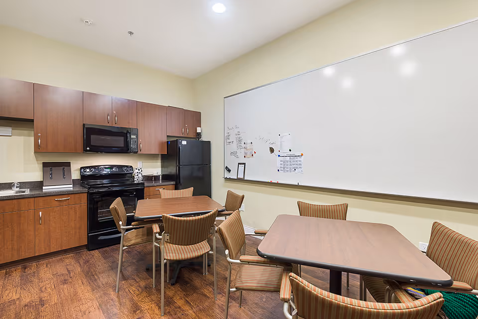 A kitchen and dining area with wooden cabinets, a black stove, microwave, and refrigerator. There are two tables with several striped chairs around them. A large whiteboard with papers and notes is mounted on the wall.
