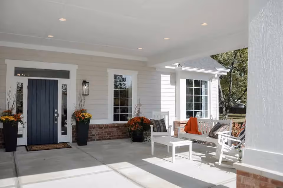 Covered front porch area of a building with a dark blue front door, two tall planters with orange and yellow flowers on either side of the door, white framed windows, white outdoor furniture including a bench and chairs with cushions, and a small white table. The porch has a concrete floor and is partially shaded.