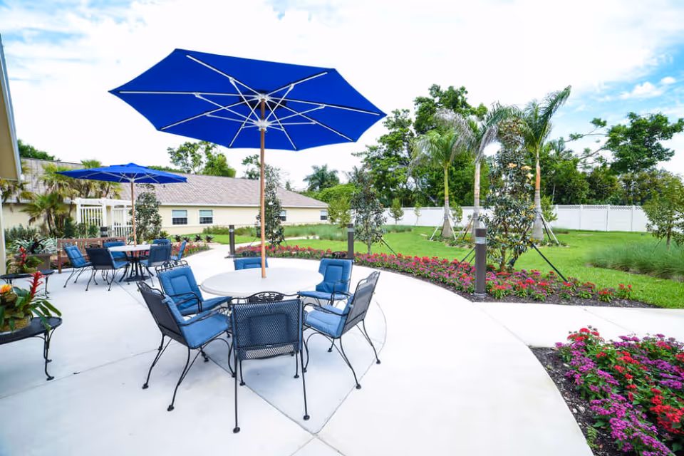 Outdoor patio area with round tables and blue cushioned chairs under large blue umbrellas, surrounded by landscaped garden beds with flowers and palm trees, adjacent to a building.