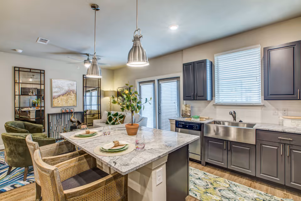 Modern kitchen and living area with a marble countertop island set for two, dark gray cabinets, stainless steel farmhouse sink, two pendant lights, green armchairs, a sofa with green pillows, decorative mirrors, and a potted plant on the island.