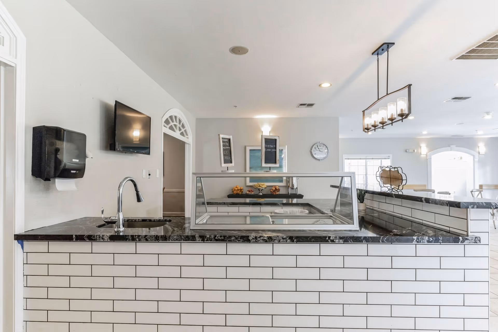 Interior view of a clean and modern serving counter area with a black marble countertop and white subway tile front. The counter has a glass sneeze guard, a sink with a faucet, a wall-mounted TV, and a paper towel dispenser. In the background, there are two small chalkboards on stands, a clock on the wall, and a table with fruit bowls. The space is well-lit with ceiling lights and a hanging light fixture.