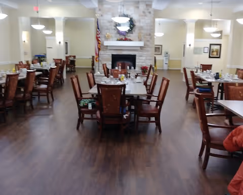 A spacious dining room with multiple wooden tables and chairs arranged neatly on a wooden floor. The room features a stone fireplace with a mantle decorated with flowers and an American flag beside it. The walls are light-colored with framed pictures and several ceiling lights providing illumination.