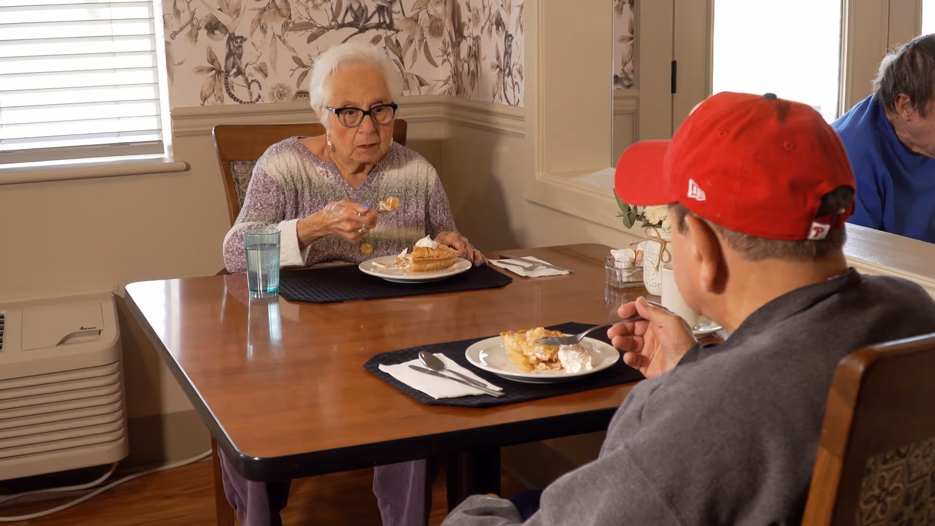 Two elderly individuals sitting at a wooden dining table eating pie with whipped cream. The woman facing the camera wears glasses and a purple sweater, while the man with his back to the camera wears a red baseball cap and a gray sweater. The room has floral wallpaper and a window with blinds.