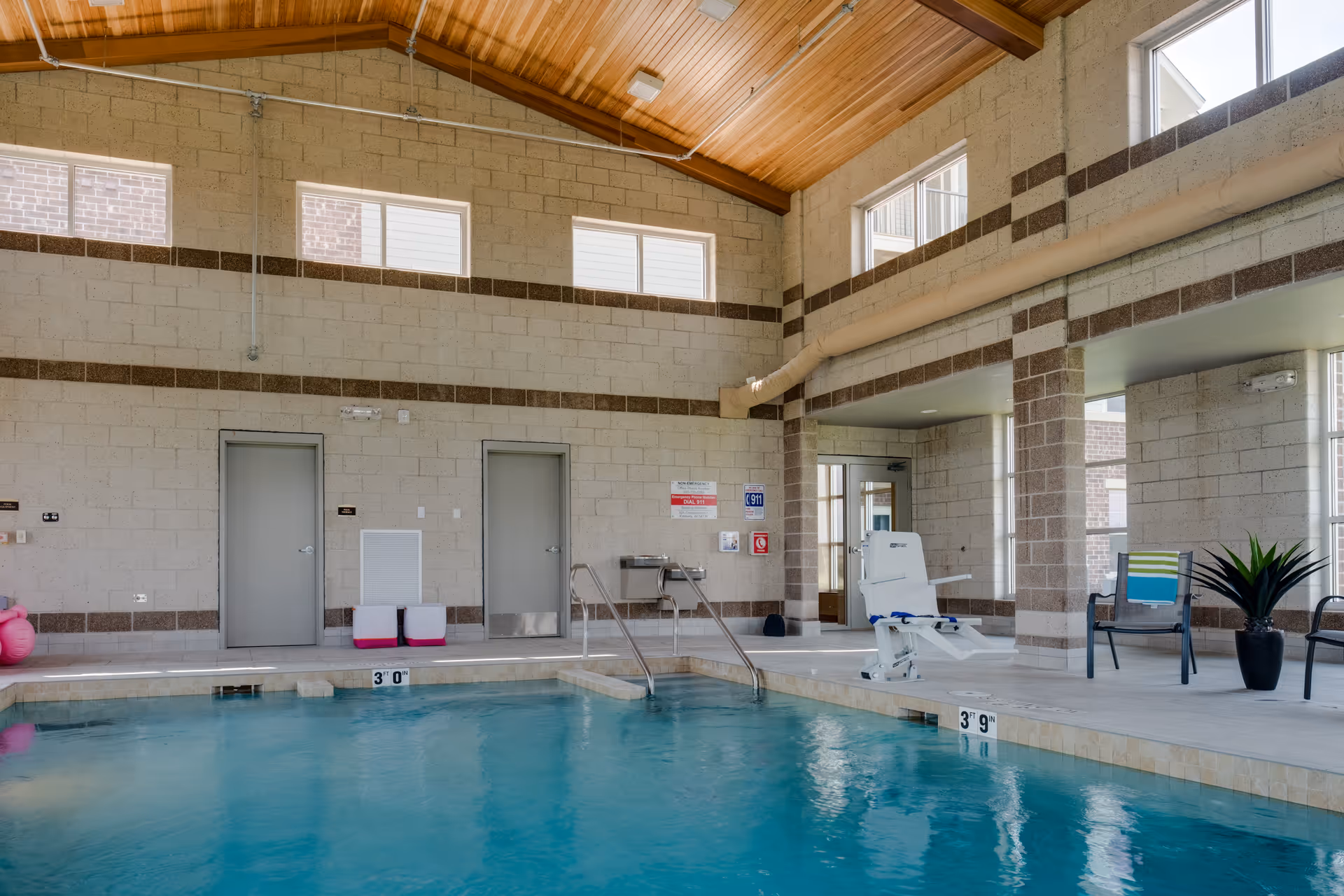 Indoor swimming pool area with clear blue water, beige tiled edges, and a pool lift chair for accessibility. The room has high ceilings with wooden paneling and multiple windows letting in natural light. There are two closed doors on one wall, a couple of chairs with a small table, and a potted plant near the poolside.