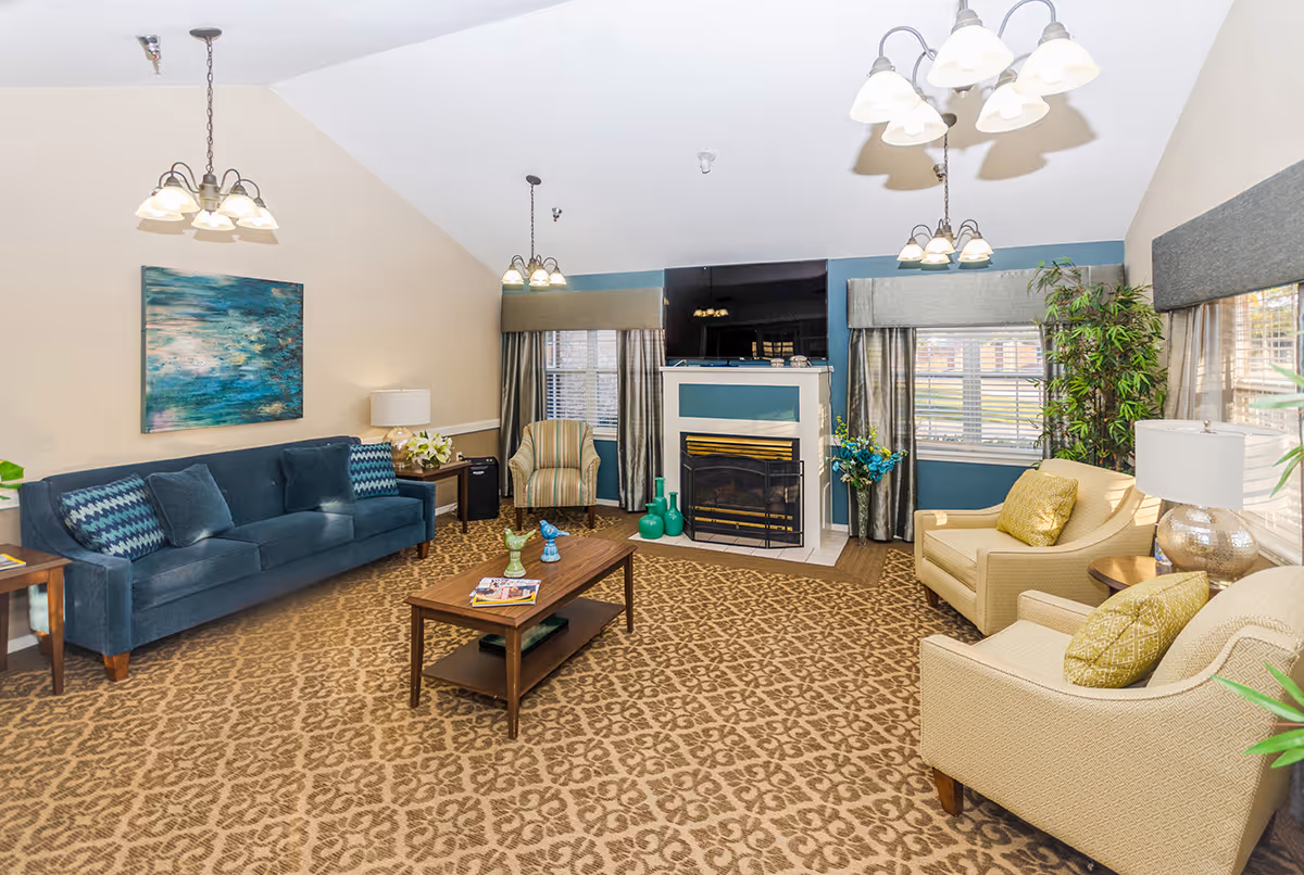 A cozy living room with a patterned carpet, a blue sofa with cushions, two beige armchairs with yellow pillows, a wooden coffee table with decorative items and magazines, a fireplace with green vases on the hearth, a large flat-screen TV mounted above the fireplace, and windows with curtains allowing natural light in. The walls are painted beige and blue, and there are ceiling light fixtures and plants adding to the decor.