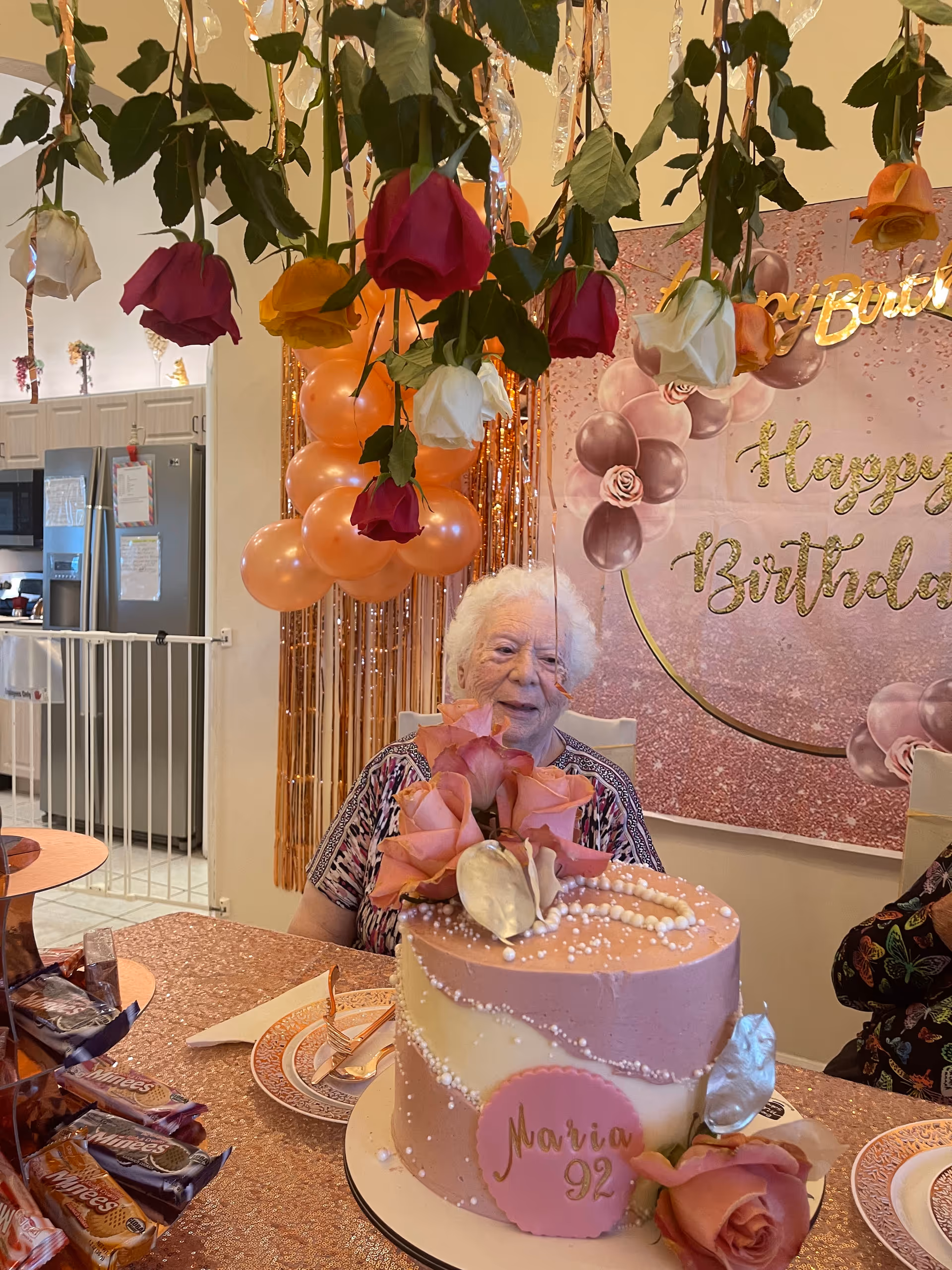 An elderly woman sitting at a decorated table with a pink and white birthday cake that says 'Maria 92'. The table is covered with a glittery rose gold tablecloth and has plates, utensils, and wrapped candies. Above the table, roses hang upside down as decoration. Behind the woman, there is a pink and gold 'Happy Birthday' banner with balloons and a kitchen area visible in the background.