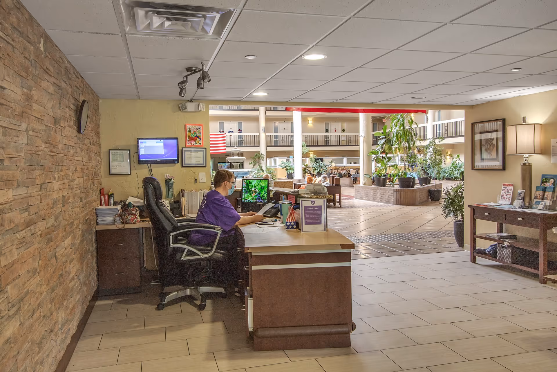 A receptionist wearing a purple shirt and face mask sits at a desk with a computer and phone in a well-lit lobby area. The lobby features a stone accent wall, tiled floor, potted plants, and seating areas with tables and chairs. The space is open with multiple floors visible in the background.
