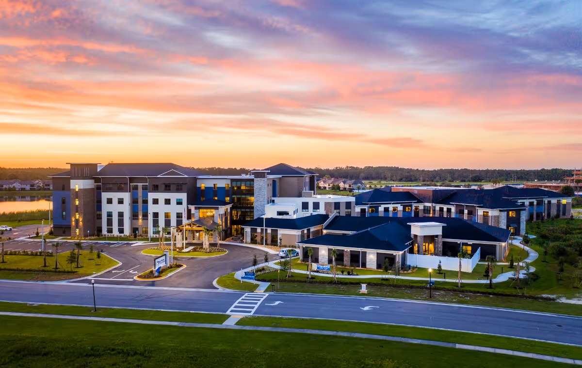 Aerial view of Blue Heron Senior Living facility at sunset, showing multiple modern buildings with well-maintained landscaping, parking areas, and a road in the foreground under a colorful sky.
