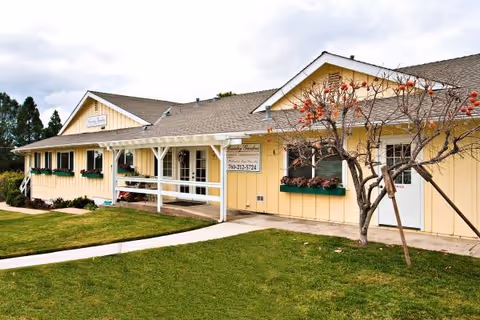 Exterior view of a yellow Country Gardens senior living building with a porch and trees.
