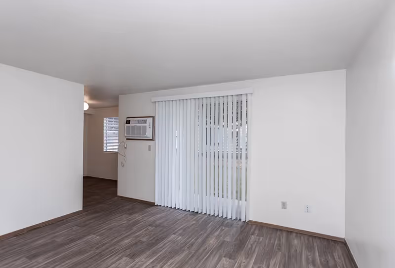 Empty living room with wood-look flooring, vertical blinds over a sliding glass door, and a wall air-conditioning unit.