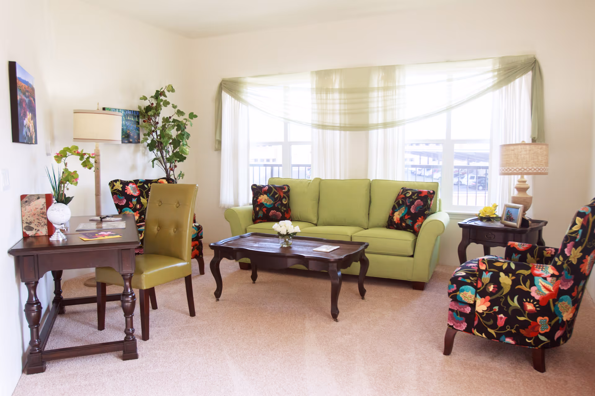 A bright living room with a light green sofa adorned with floral patterned pillows, two matching floral armchairs, a dark wooden coffee table with a small flower arrangement, a side table with a lamp and picture frame, a small desk with a green chair, and a large window with sheer curtains and a green swag valance.