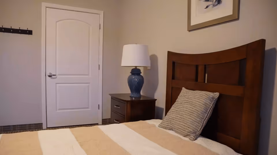 A bedroom with a wooden headboard bed, a striped pillow, and a beige and white striped blanket. Next to the bed is a wooden nightstand with a blue ceramic lamp. A closed white door and a wall-mounted coat rack with hooks are visible in the background.