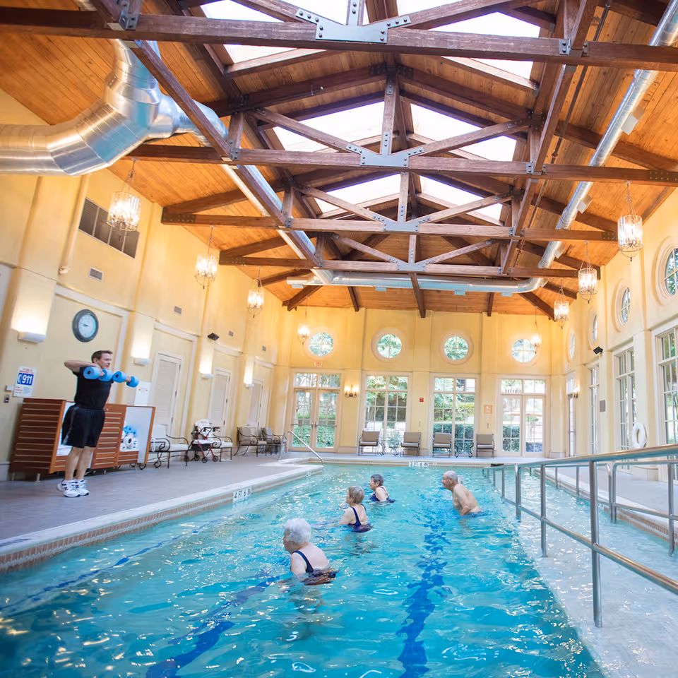 Indoor swimming pool with four elderly people participating in a water exercise class led by an instructor holding blue foam dumbbells. The room features a high wooden ceiling with exposed beams and large windows letting in natural light.