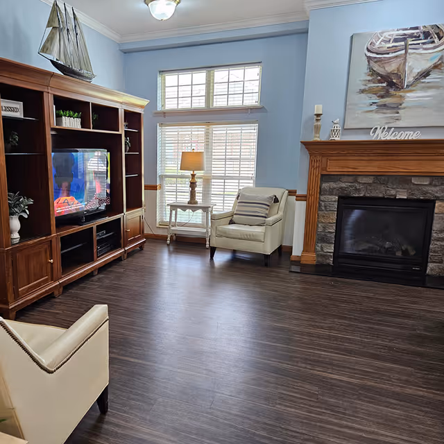 Cozy living room with a wooden entertainment center and TV, two armchairs by a window, a lamp, and a stone fireplace.