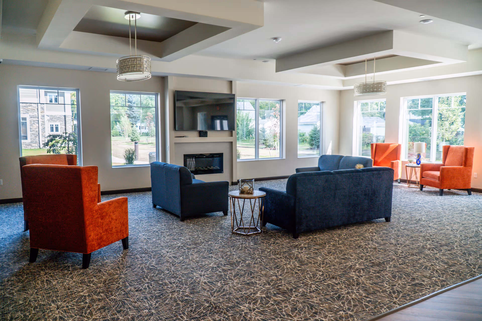 A bright and spacious living room area with large windows letting in natural light. The room features two blue sofas, two orange armchairs, a small round table, a wall-mounted flat-screen TV above a modern fireplace, and patterned carpet flooring.