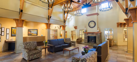 A spacious living room area with high vaulted wooden ceilings and large chandeliers. The room features a stone fireplace with a clock above it, surrounded by beige walls and wooden beams. There are multiple seating options including patterned armchairs and dark blue sofas arranged around wooden coffee tables. The room is well-lit with natural light coming through windows and has a warm, inviting atmosphere.