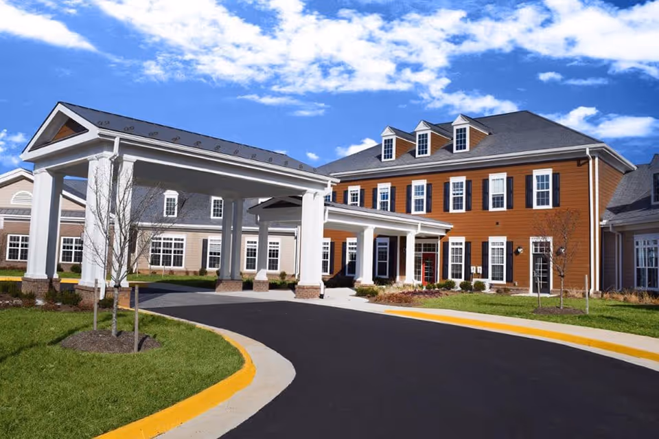 Exterior view of a senior living facility building with a covered entrance driveway, manicured lawn, and clear blue sky with some clouds.