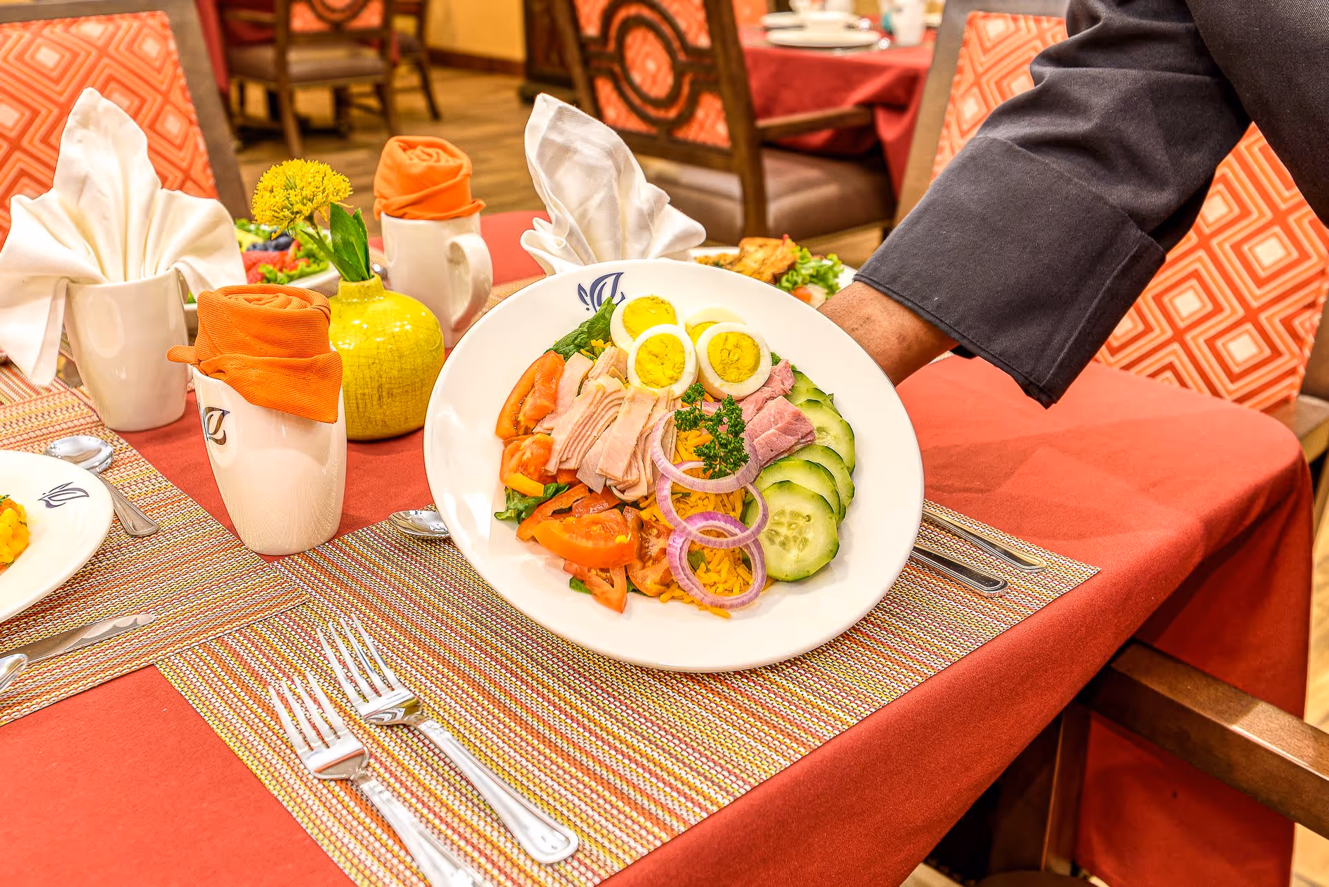 A hand is serving a plate of salad with sliced boiled eggs, tomatoes, cucumbers, ham, and onions on a table set with orange and white napkins, silverware, and a small yellow vase with a flower in a dining room.