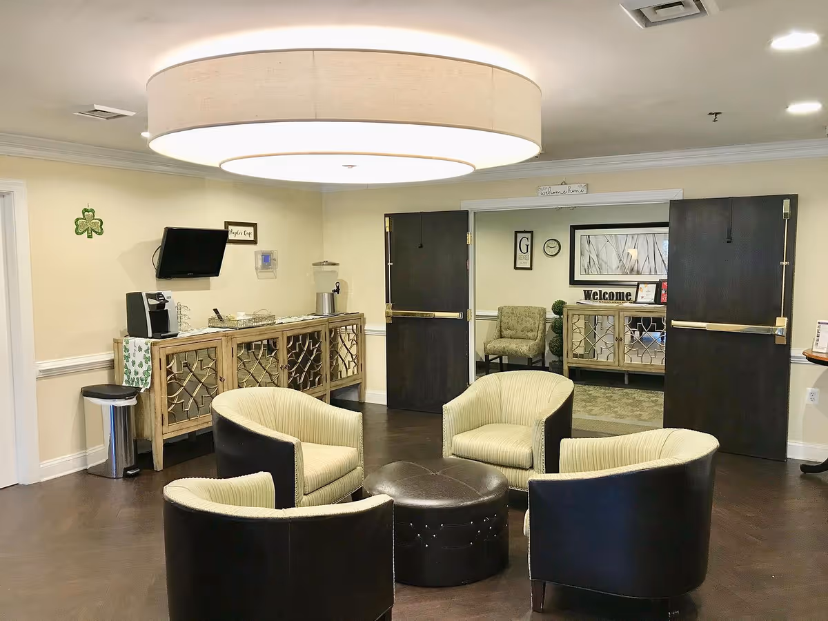 A cozy seating area in a senior living facility with four cushioned armchairs arranged around a round ottoman. Behind the seating area is a wooden sideboard with a coffee machine, water dispenser, and some cups. The room has beige walls, dark wooden double doors, and a large circular ceiling light fixture. A small TV is mounted on the wall above the sideboard, and there is a shamrock decoration on the wall.