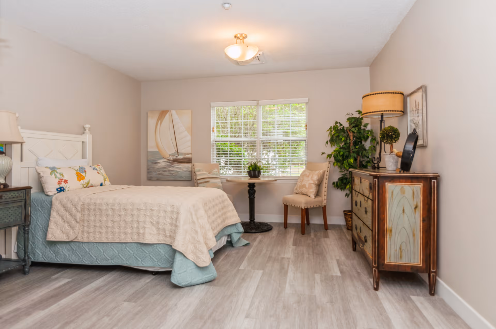 Bright furnished bedroom with a bed, nightstand, dresser, chair and a window overlooking greenery.