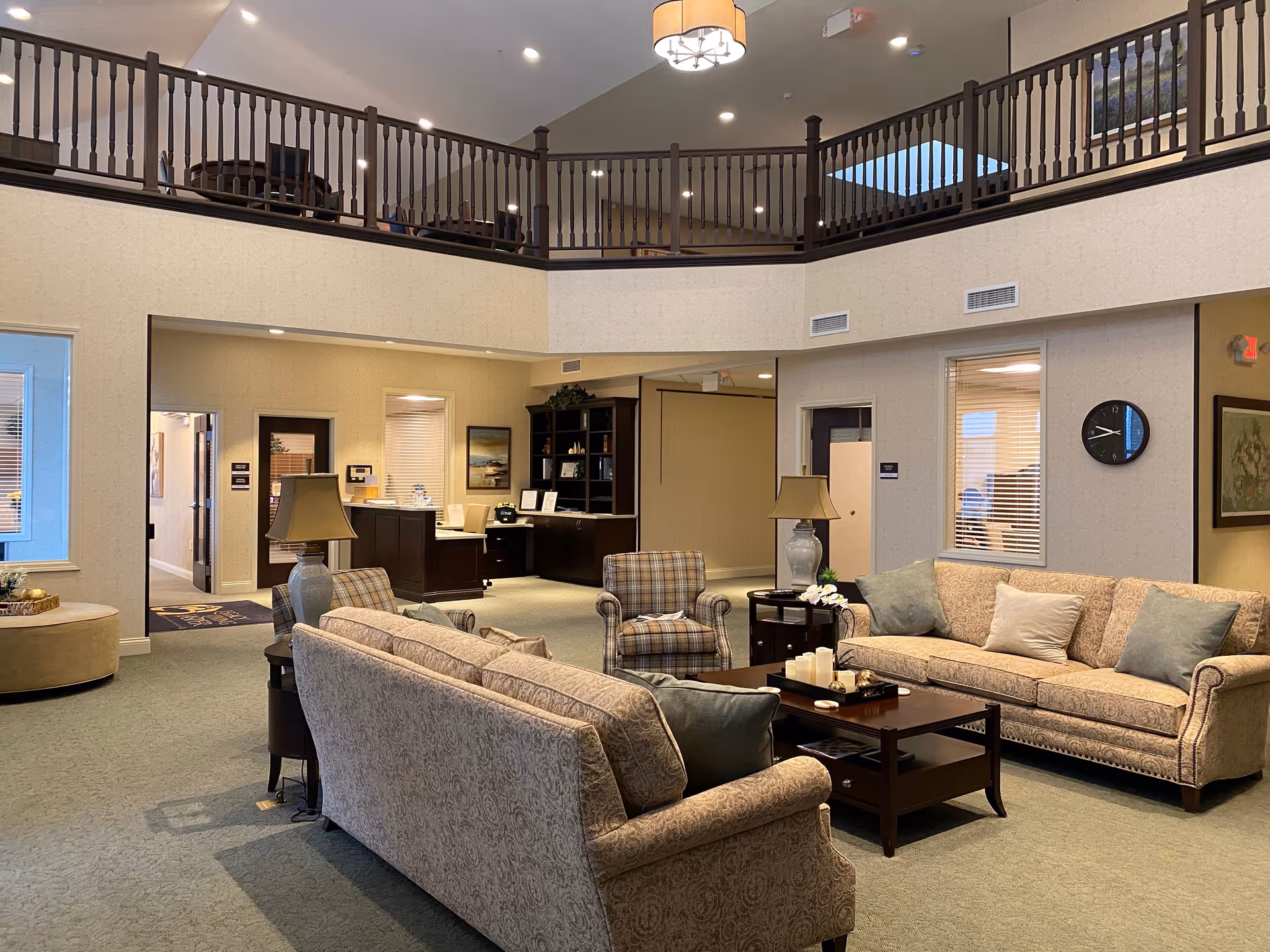 A spacious and well-lit living room area in Legacy Living Jasper featuring two patterned sofas, two armchairs, a dark wood coffee table with candles, and two table lamps. The room has a high ceiling with a balcony railing above and neutral-colored walls and carpet. There is a clock on the wall and an office area visible in the background.