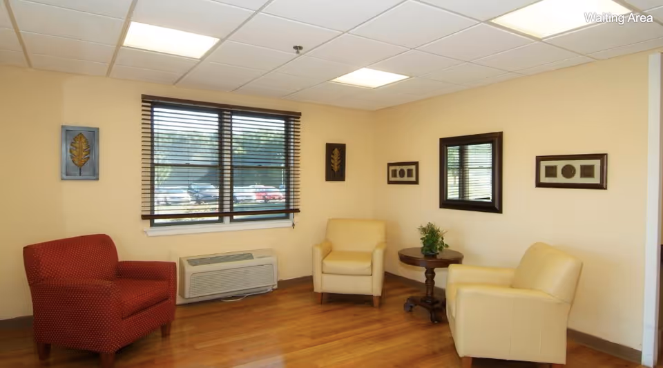 A bright waiting room with three armchairs around a small table, wall art, a window with blinds, and hardwood floor.