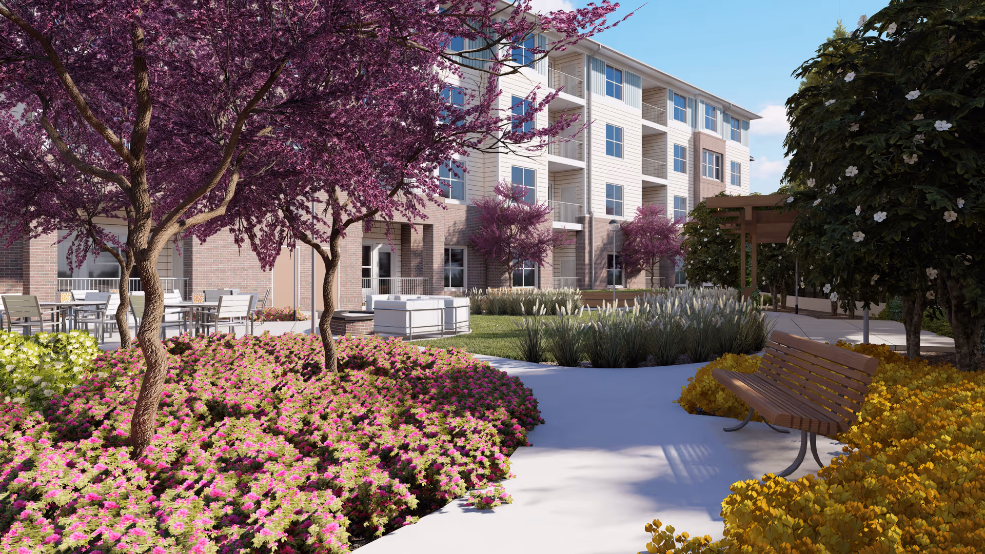 Outdoor garden area at Acclaim at Cary Pointe featuring a paved walkway, wooden bench, flowering trees with purple blossoms, colorful flower beds, and a multi-story residential building in the background under a clear blue sky.