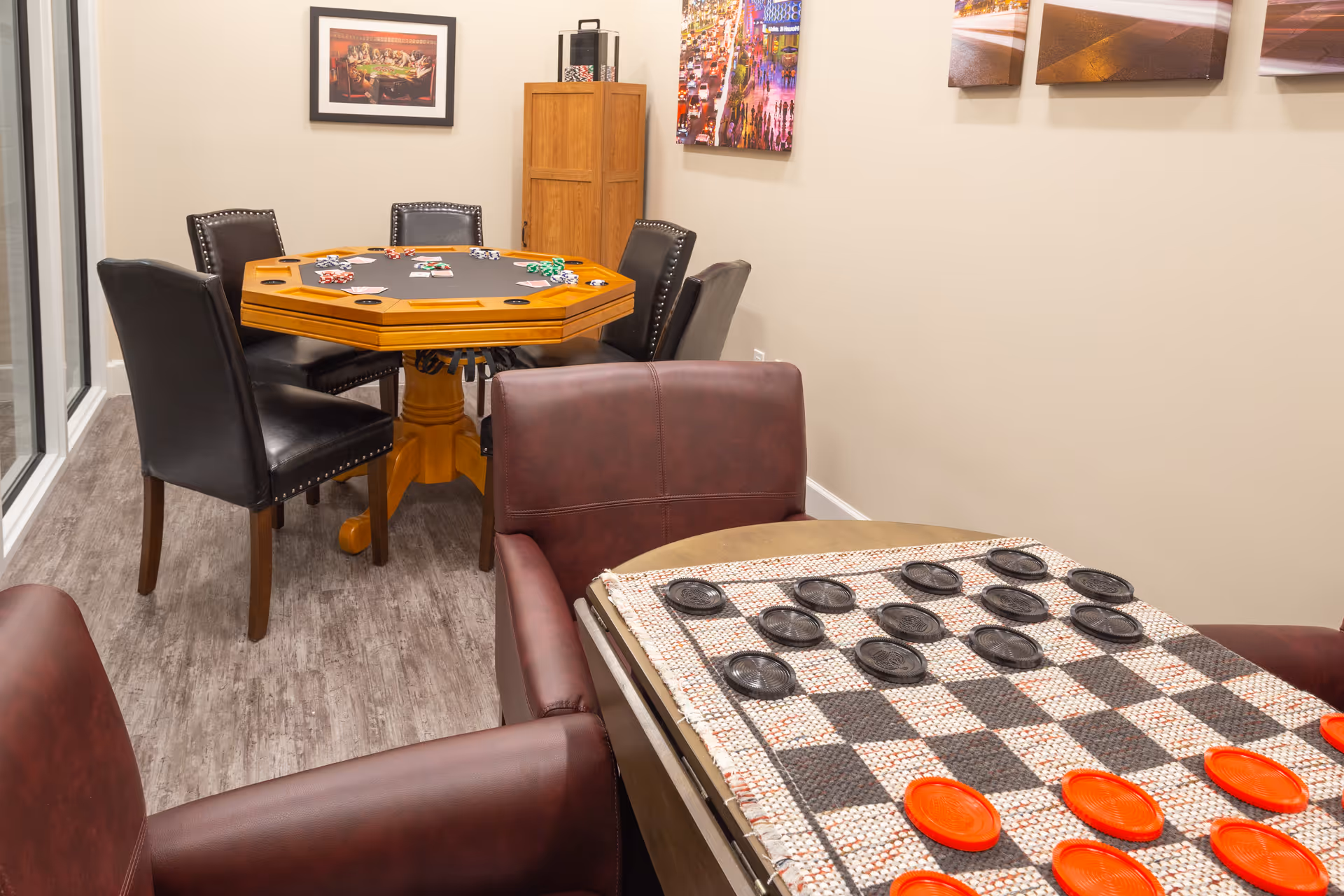 A cozy game room featuring a wooden octagonal poker table with black leather chairs and poker chips, and a checkers board with red and black pieces on a small table surrounded by brown leather chairs. The room has light-colored walls adorned with framed pictures and wood flooring.