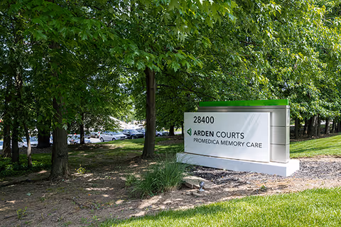 Entrance sign reading '28400 Arden Courts ProMedica Memory Care' on a grassy lawn with trees and parked cars in the background.
