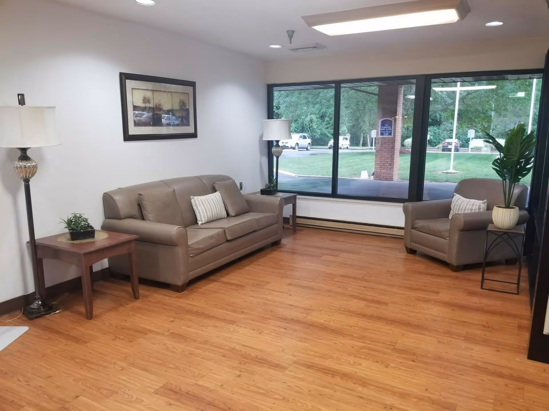 A waiting area with a brown leather sofa and matching armchair, both with decorative pillows. There are two side tables, one with a small plant and the other with a lamp. A large window shows a view of the outside greenery and parked cars. The floor is wooden, and a framed picture hangs on the white wall.