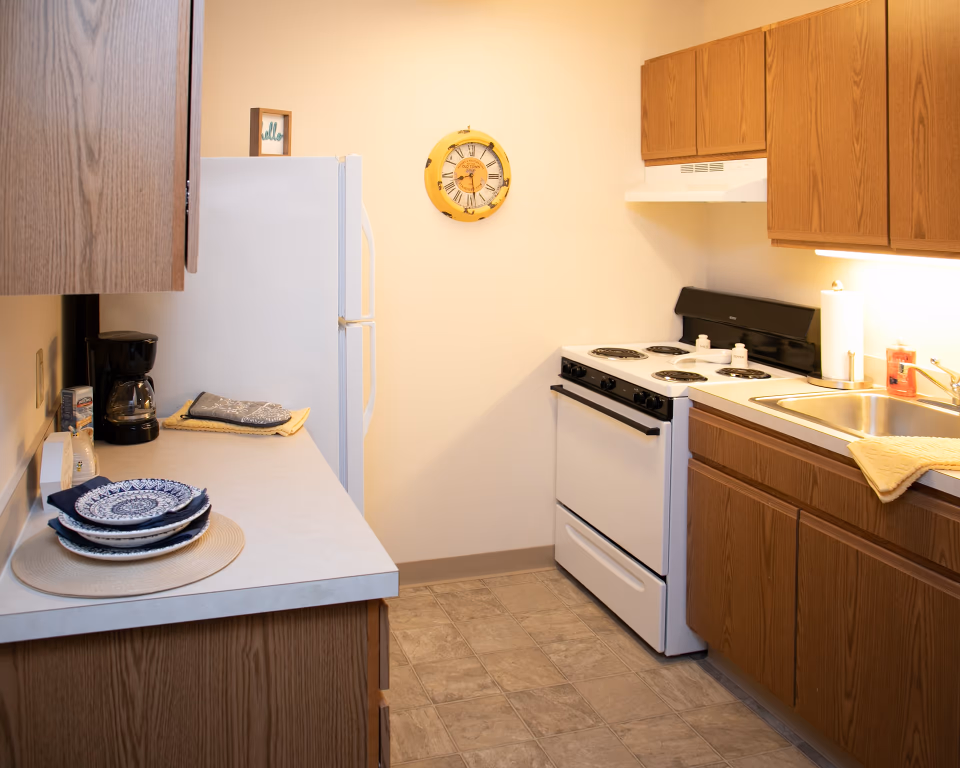A small kitchen with wooden cabinets, a white refrigerator, a white electric stove, a stainless steel sink, and a countertop with a coffee maker, plates, and kitchen towels. A yellow clock hangs on the wall above the stove.