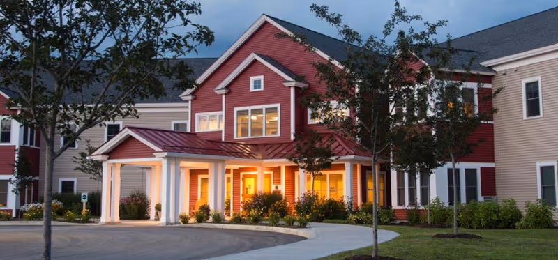 Front exterior of a red and beige senior living building with a covered, illuminated entrance, landscaped grounds, and a curved driveway.