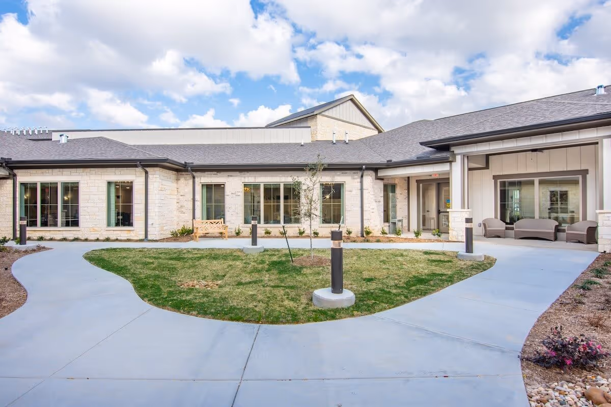 Exterior view of StoneCreek of Copperfield Senior Living facility showing a single-story building with light-colored stone walls, large windows, and a covered seating area with outdoor furniture. There is a curved concrete walkway surrounding a small grassy area with young trees and modern outdoor lights under a partly cloudy sky.