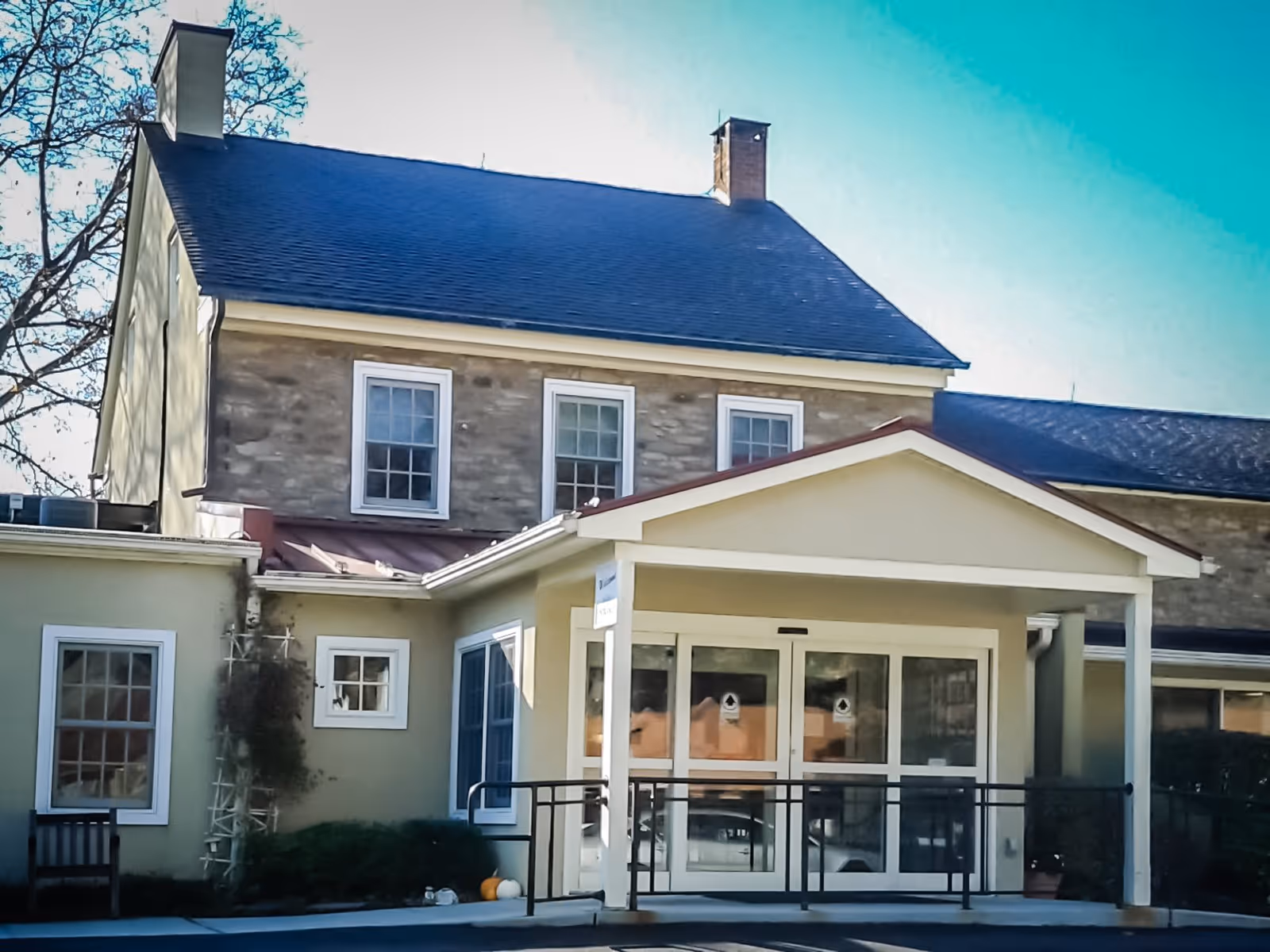 Exterior view of a two-story building with a stone facade on the upper floor and light-colored walls on the lower floor. The entrance has a covered porch with a ramp and double glass doors. There are several windows and some greenery near the entrance.