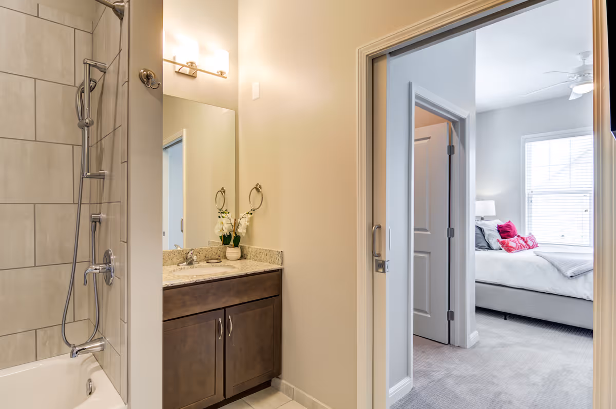View of a bathroom with a bathtub and shower combination on the left, a vanity with a sink and mirror in the center, and an open doorway leading to a bedroom with a bed, pillows, and a window with blinds on the right.