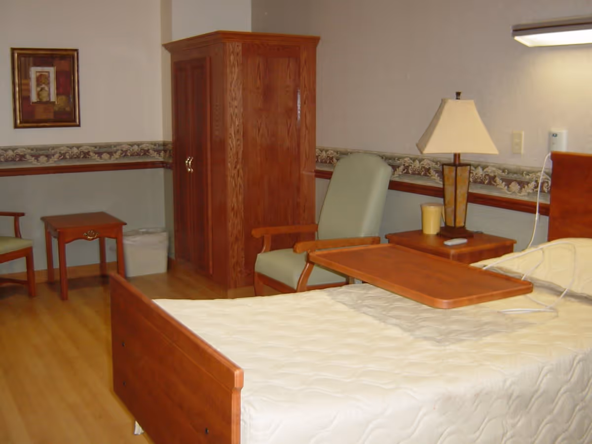 A senior living facility bedroom with a wooden bed featuring a white quilted bedspread and an overbed table. There is a wooden nightstand with a lamp and a cup, a green cushioned armchair, a wooden armoire, a small wooden side table, and another chair. The room has light-colored walls with a decorative border and wood trim, and a light wood floor.