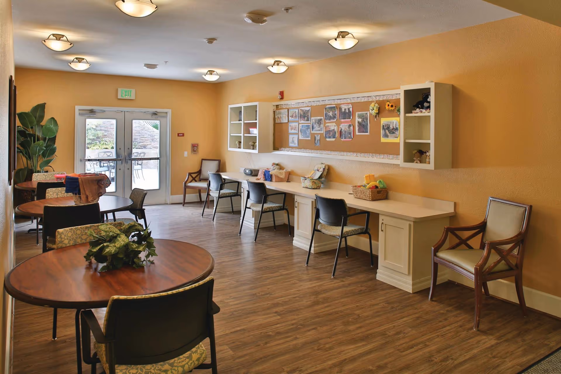 A well-lit common area with round wooden tables and chairs, a long counter with chairs against a wall displaying photos and decorations, and double glass doors leading to an outdoor patio.