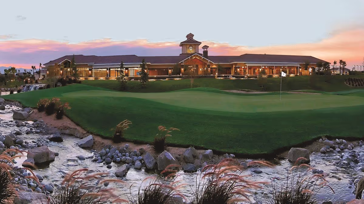 A scenic view of a golf course with a putting green and a flag, a rocky stream in the foreground, and a large clubhouse building with a clock tower in the background during sunset.