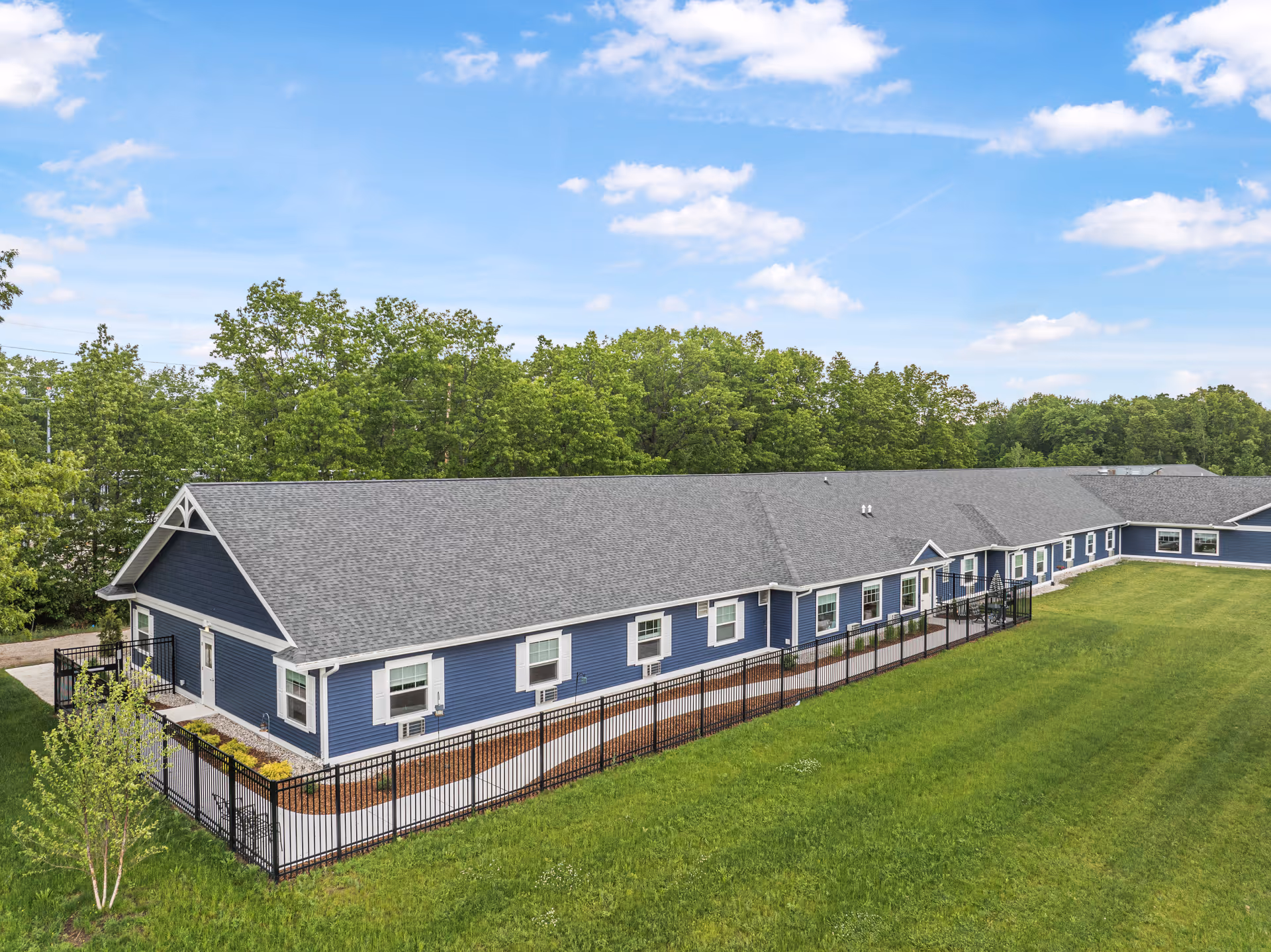 Aerial view of a single-story blue senior living building with a fenced lawn and trees in the background.
