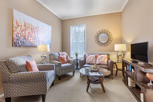 Cozy living room with patterned armchairs, a small sofa, glass coffee table, TV console, and wall art.