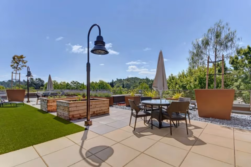 Outdoor patio area with a round table and four chairs under a closed umbrella, large potted plants, raised wooden garden beds, green artificial turf, and a tall streetlamp under a clear blue sky.