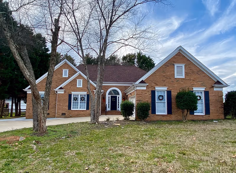 Brick ranch-style house with an arched central entry, blue shutters, and leafless trees on a grassy front lawn under a cloudy sky.