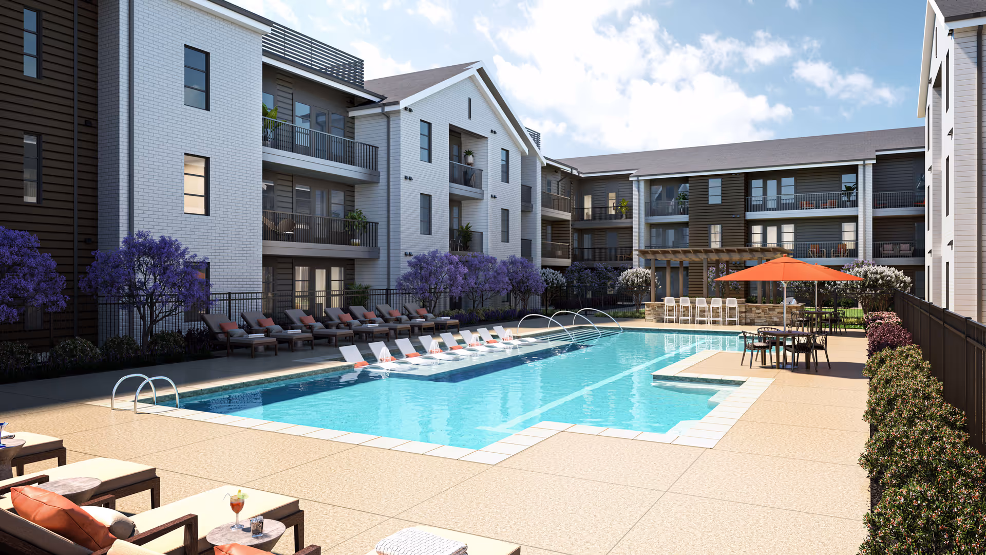Outdoor swimming pool area surrounded by lounge chairs with orange cushions, tables with umbrellas, and a bar seating area under a pergola. The pool is located in the courtyard of a multi-story residential building with balconies and purple flowering trees along the perimeter.